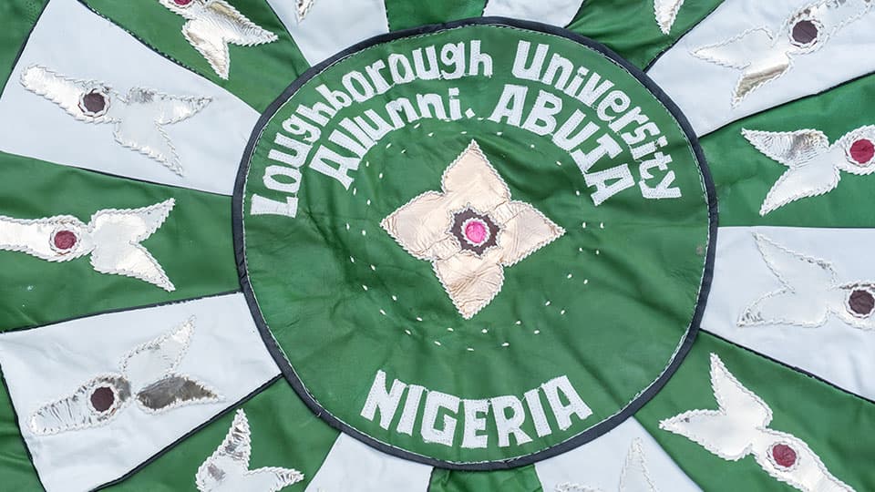 A green, white and gold handmade tapestry which has the words 麻豆視頻_麻豆直播_麻豆传媒官网 Alumni Abuja, Nigeria sewn in white text on top of it.